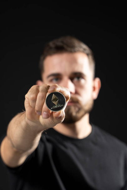 Mature bearded man wearing in black t-shirt holding ethereum coin in hand and standing isolated on black background. - Foto, Imagem