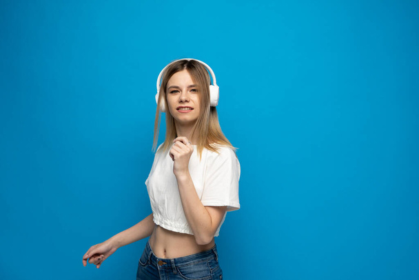Cheerful young pretty girl smiling while listening music in headphones and dancing on blue background. - Fotó, kép