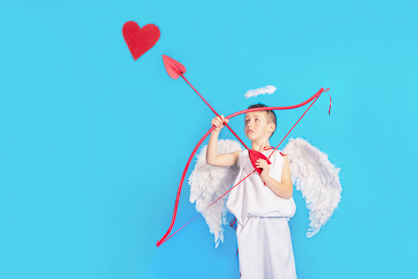 Little Boy in een cupido kostuum met vleugels en een halo houdt een pijl en boog, richt zich op het hart, selectieve focus. Trieste cupido schiet op het hart. Wenskaart por Valentijnsdag, - Foto, afbeelding