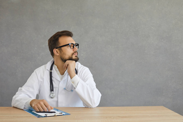 Dreamful pensive male doctor in white medical gown suit sit at desk look aside - Photo, Image