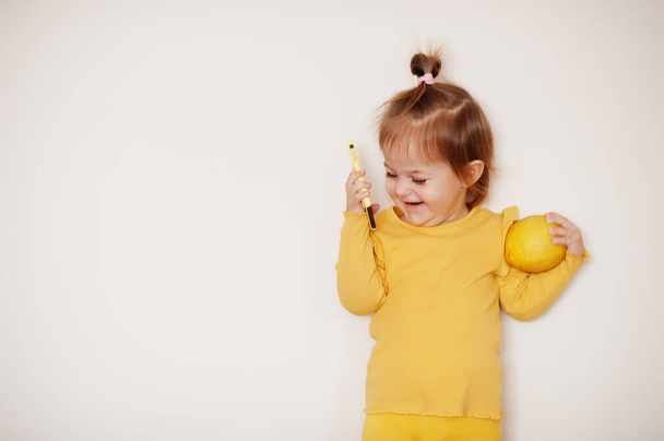 Baby girl in yellow with lemon and mobile phone, isolated background. - Foto, imagen