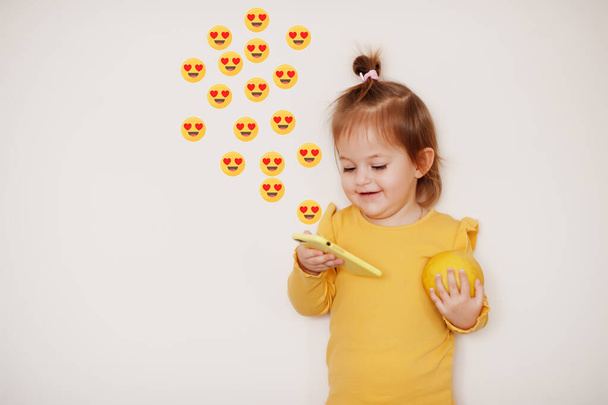 Baby girl in yellow with lemon and mobile phone with heart eyes emoji, isolated background. - Foto, Bild