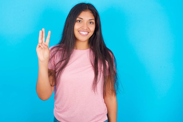 Девушка-подросток показывает пальцем номер три, улыбаясь уверенно и счастливо. Молодая красивая латиноамериканская брюнетка в розовой футболке позирует на синем фоне - Фото, изображение