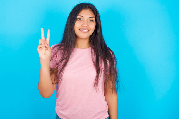 10代の女の子は自信と幸せに微笑んでいる間,指のナンバー2を指さして指を指さす. 青い背景にポーズピンクTシャツを着ている若い美しいヒスパニックブルネット女性 - 写真・画像