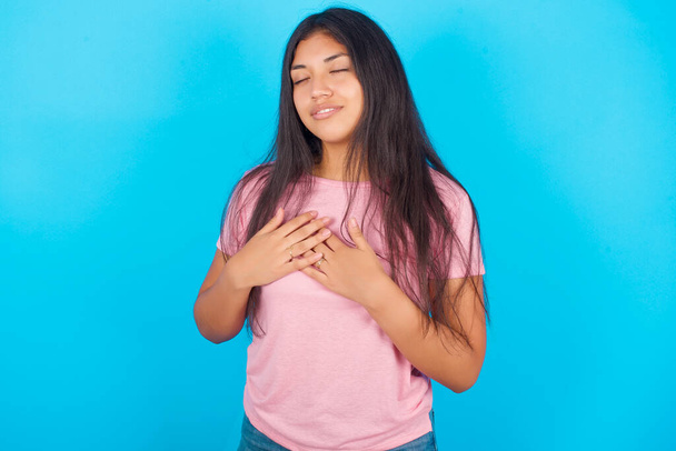 дівчина-підліток посміхається руками на грудях із закритими очима і вдячним жестом на обличчі. Концепція здоров'я. Молода красива іспаномовна брюнетка в рожевій футболці позує на синьому фоні - Фото, зображення