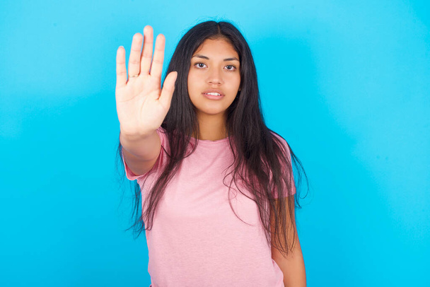 Девочка-подросток прекращает петь с ладонью на руке. Предупреждающее выражение с негативным и серьезным жестом на лице. Молодая красивая латиноамериканская брюнетка в розовой футболке позирует на синем фоне - Фото, изображение
