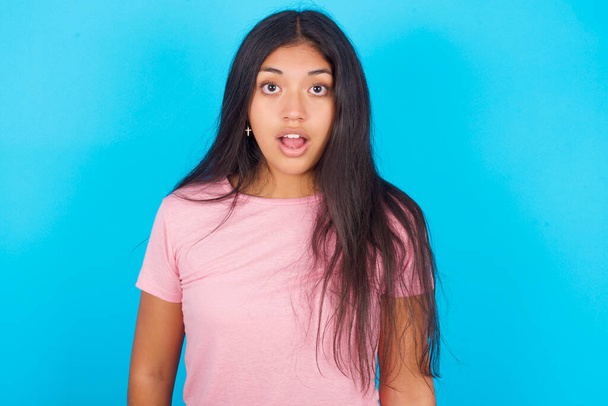 Oh my God. Surprised  girl  stares at camera with shocked expression exclaims with unexpectedness. Young beautiful Hispanic brunette woman wearing pink T-shirt posing against blue background - Photo, Image