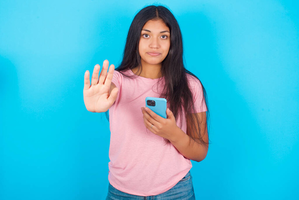  κορίτσι που χρησιμοποιεί και γραπτών μηνυμάτων με smartphone με ανοικτό χέρι κάνει σήμα στοπ με σοβαρή και σίγουρη έκφραση, χειρονομία άμυνας. Νέα όμορφη ισπανόφωνη μελαχρινή γυναίκα φοράει ροζ T-shirt ποζάροντας σε μπλε φόντο - Φωτογραφία, εικόνα