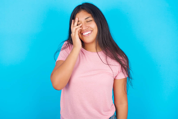  girl makes face palm and smiles broadly, giggles positively hears funny joke poses. Young beautiful Hispanic brunette woman wearing pink T-shirt posing against blue background - Photo, Image
