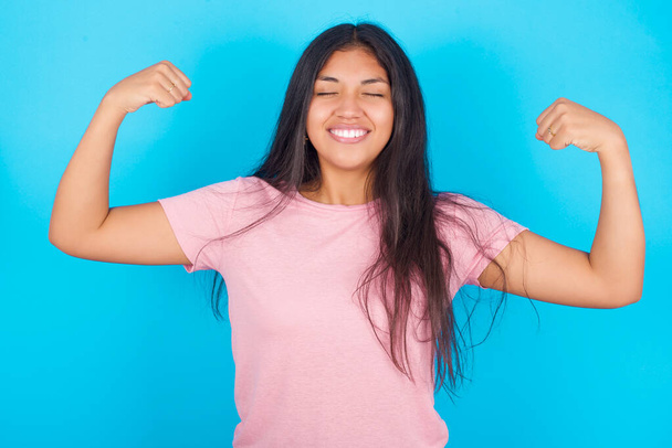 Strong powerful  woman  toothy smile, raises arms and shows biceps. Look at my muscles! Young beautiful Hispanic brunette woman wearing pink T-shirt posing against blue background - Photo, Image