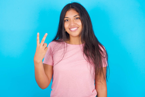  nő mosolyog és barátságos, mutatja a második vagy második kéz előre, visszaszámlálás. Fiatal gyönyörű spanyol barna nő visel rózsaszín póló pózol ellen kék háttér - Fotó, kép