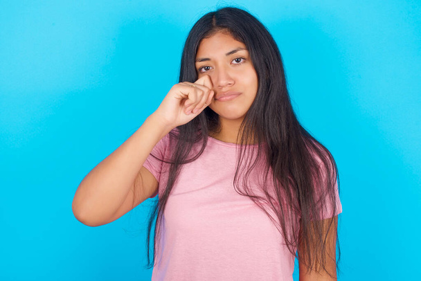 Грустная молодая красивая латиноамериканская брюнетка в розовой футболке на синем фоне вытирает слезу со щеки - Фото, изображение