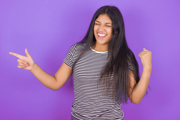Fröhliche junge Lateinerin in gestreiftem T-Shirt vor lila Hintergrund zeigt Kopierraum und feiert Glück - Foto, Bild