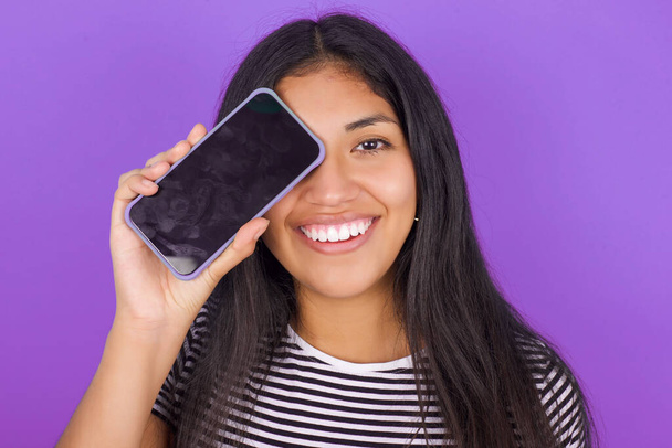 nuori latino nainen yllään raidallinen T-paita tilalla moderni älypuhelin peittää toisen silmän hymyillen - Valokuva, kuva