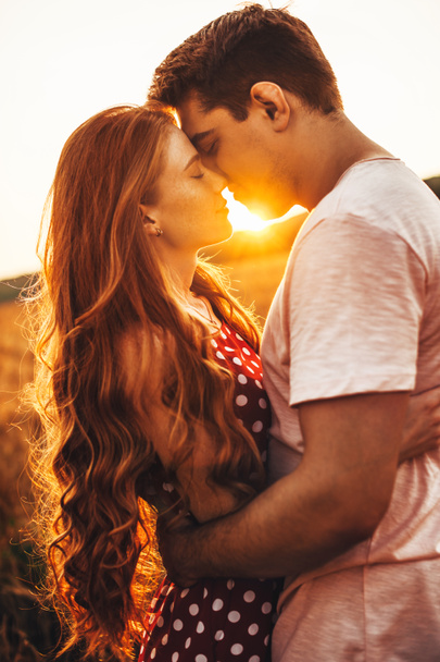 Vista lateral de una mujer de pelo largo y rojo besando a su marido en una reunión al aire libre. Posando sobre el fondo brillante - Foto, Imagen