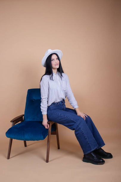 attractive brunette woman in blouse, hat and jeans on a blue chair on brown background. female fashion portrait - Foto, afbeelding