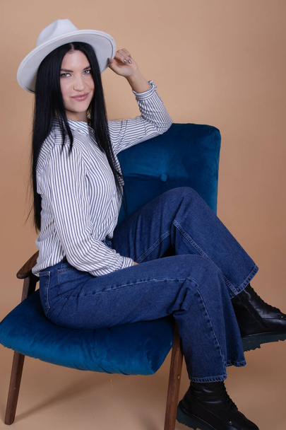 portrait of young woman in white blouse, white hat and jeans near blue chair on brown background. pretty female portrait - Fotó, kép