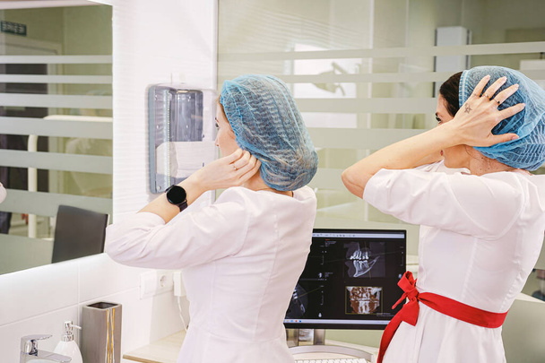 Dos atractivas mujeres dentistas preparándose para el trabajo poniendo gorras médicas y mirando el espejo en la clínica dental. - Foto, Imagen