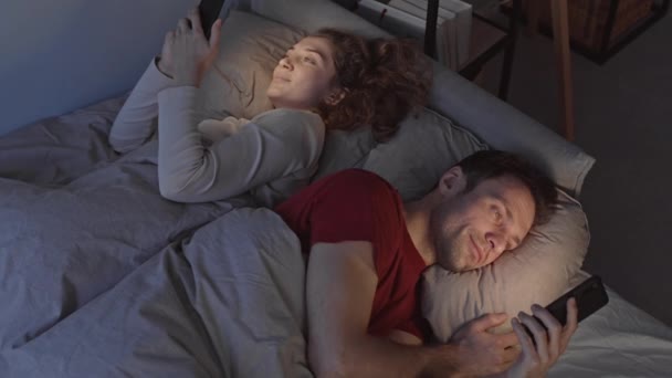 睡眠に行く前に、彼らのスマートフォンを使用して若いカップルのトップビューの遅いショット,夜遅くにベッドで一緒に横たわっています - 映像、動画
