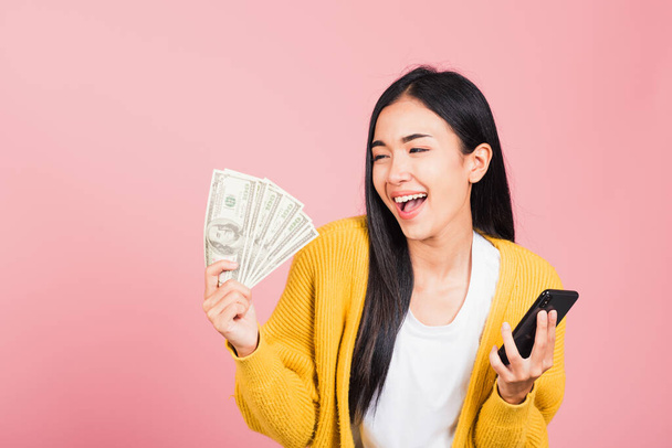 Retrato asiático feliz hermosa joven mujer adolescente shopper sonriendo de pie emocionado sosteniendo en línea teléfono móvil inteligente y billetes de dinero en dólares en la mano en verano, estudio de tiro aislado sobre fondo rosa - Foto, imagen