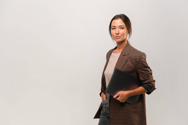 Elegante mujer asiática confiada de pie con el ordenador portátil en las manos y mirando a la cámara en el fondo gris. Retrato de mujer sofisticada - Foto, Imagen
