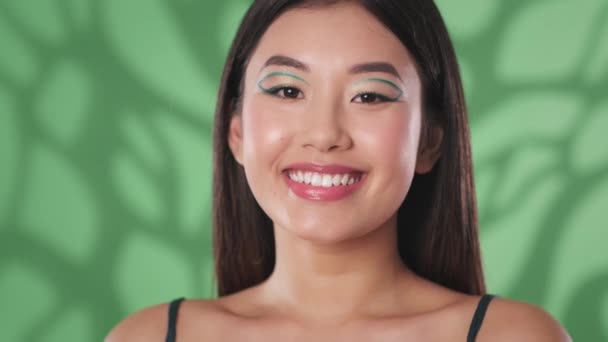 Maquillage artistique créatif. Jeune belle femme asiatique avec des flèches vertes sur les yeux souriant à la caméra, fond de studio vert - Séquence, vidéo