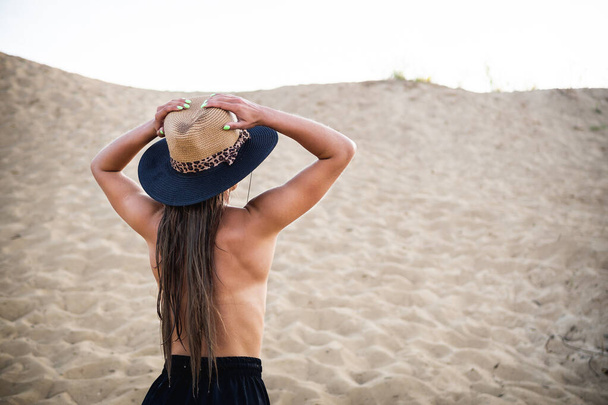 nacktes brünettes Model in schwarzer Hose und Hut, das im Sand vor der Kamera posiert. Nah- und Langdreh. - Foto, Bild
