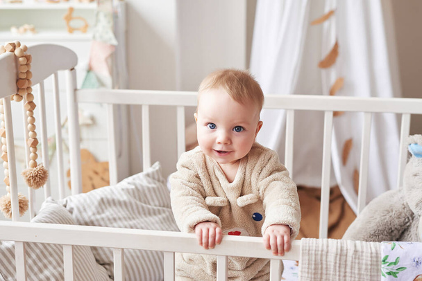 Baby 6 months standing in crib in children's room. Happy motherhood and fatherhood. Maternity hospital and clinic. Father and mother day. Early childhood development, nursery. - Foto, afbeelding