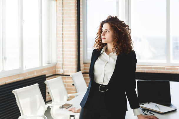 Portrait of a beautiful successful young business woman with red curly hair in the interior of the office stands leaning on the table near the window - Foto, Bild