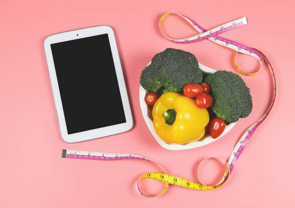 Top view or flat lay of computer tablet, vegetables capsicum, broccoli and tomato in heart shape plate with measuring tape on pink background with copy space, healthy lifestyle and weight loss concept. - Fotografie, Obrázek
