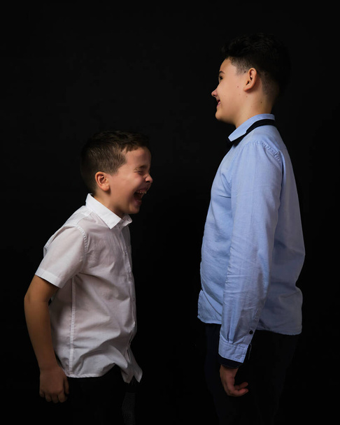 Two boys on a black background are brothers.Senior and junior.Atelier.The younger one laughs at the older brother.Open mouth - Fotografie, Obrázek