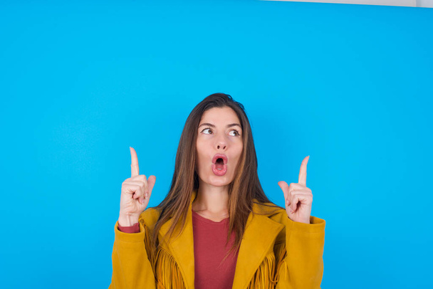  woman wearing jacket over blue studio background being amazed and surprised looking and pointing up with fingers showing something strange. - Photo, Image