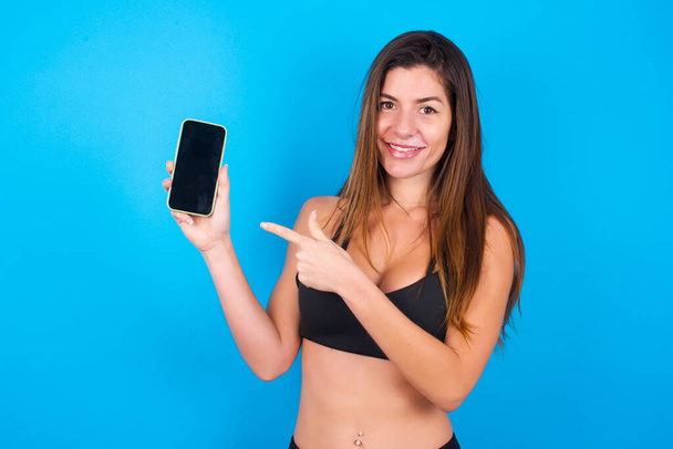 Smiling  brunette woman wearing sportswear showing and pointing at empty phone screen. Advertisement and communication concept. - Foto, Imagem