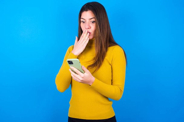 młoda piękna kobieta w żółtym swetrze nad niebieskim tle studio jest głęboko zaskoczony, gapi się na wyświetlaczu smartfona, czyta szokujące wiadomości na stronie internetowej, Omg, jego straszne! - Zdjęcie, obraz