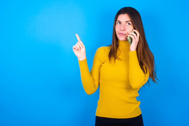 młoda piękna kobieta w żółtym swetrze nad niebieskim tle studio mówi przez telefon komórkowy spędza wolny czas w pomieszczeniach połączeń do znajomego. - Zdjęcie, obraz