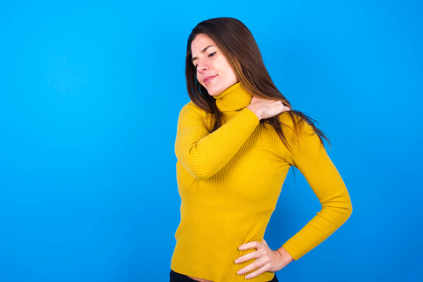 joven hermosa mujer usando amarillo suéter sobre azul fondo del estudio tiene dolor de espalda en el estudio - Foto, Imagen