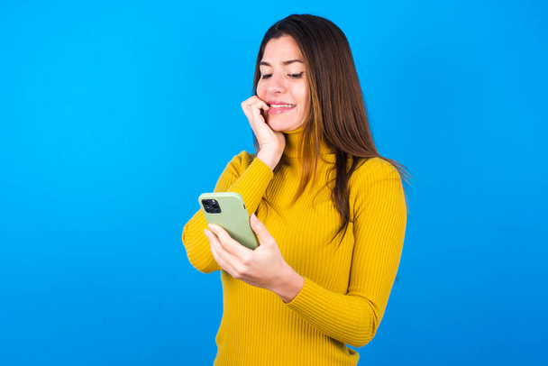 Portrait of pretty frightened young beautiful woman wearing yellow sweater over blue studio background chatting biting nails after reading some scary news on her smartphone. - Photo, image