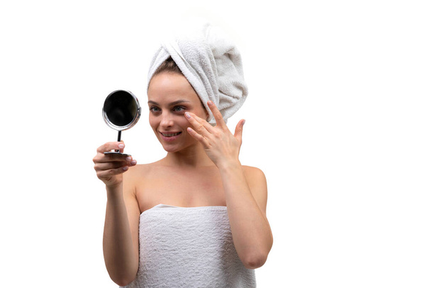 beautiful middle-aged woman with well-groomed skin applies cream to her face and looks in a small mirror on a white background - Photo, Image