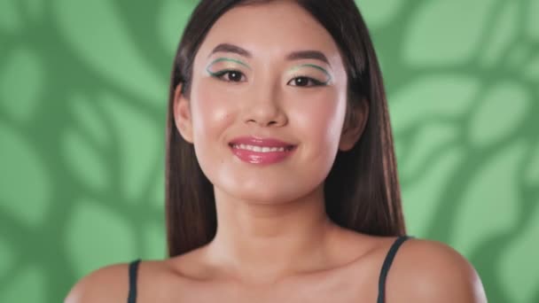 Yaratıcı makyajlı genç Asyalı bayan kameraya öpücük yolluyor ve gülümsüyor, yeşil arka planda flört ediyor. - Video, Çekim