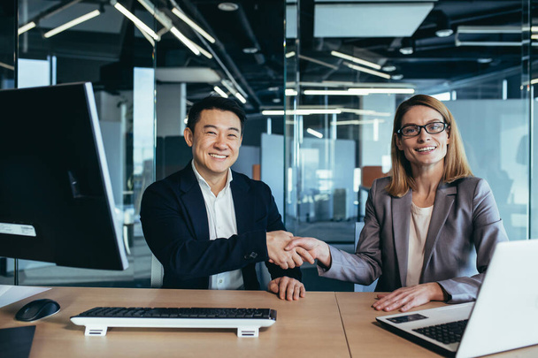 Two workers shaking hands, looking at the camera and smiling, Asian man and woman business people working in a modern office - Zdjęcie, obraz