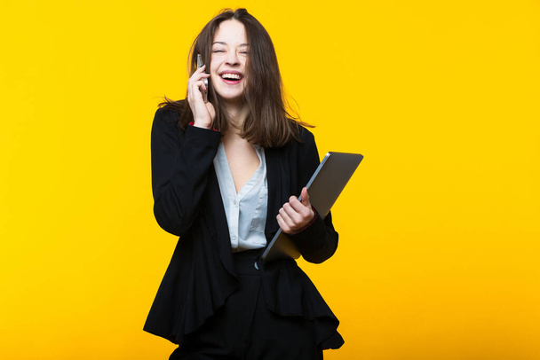 Eine junge Frau im Anzug telefoniert und hält einen Laptop vor gelbem Hintergrund - Foto, Bild