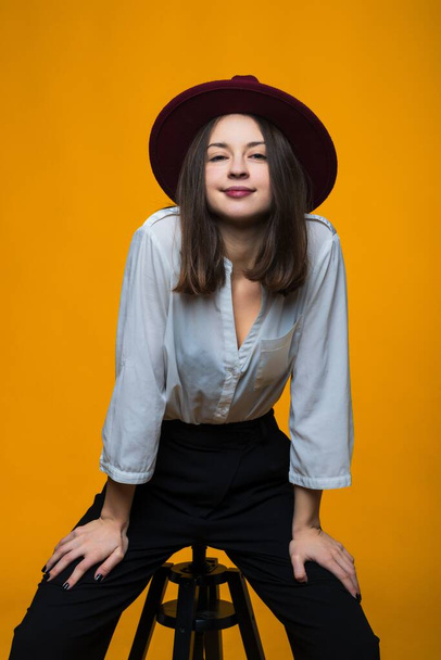 A beautiful brunette in a hat and a white shirt is sitting on a chair on a yellow background - Foto, Bild