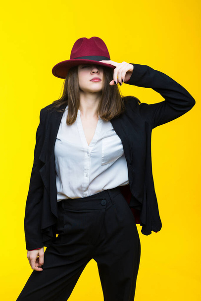 Brunette in a black suit, white shirt and hat on a yellow background - Photo, Image