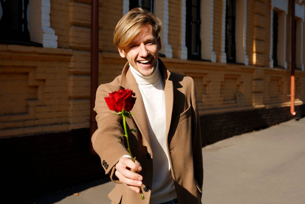 Happy handsome man smiling while giving red rose outside in street. Valentine's Day, 8th March, birthday, anniversary romantic love concept. - Foto, imagen