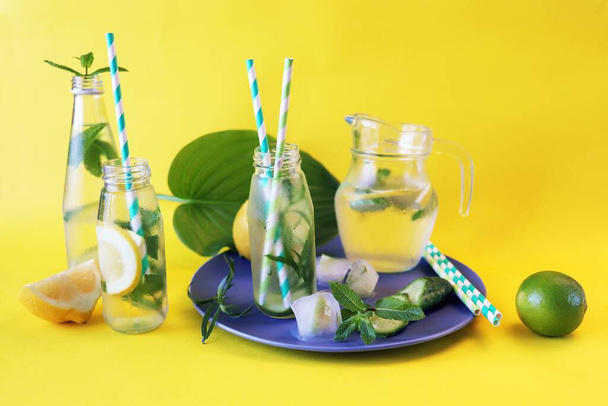 Limonlu su, dilimlenmiş salatalık, nane, tarhun ve buz, parlak sarı bir arka planda, cam şişelerde kokteyl tüpleriyle yenilenen sağlıklı bir içecek. - Fotoğraf, Görsel