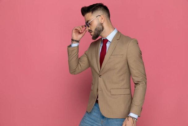 side view of a handsome businessman arranging his sunglasses and posing with attitude on pink background - Foto, afbeelding