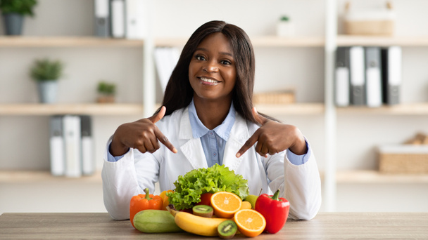 Muotokuva onnellinen afrikkalainen amerikkalainen ravitsemusterapeutin osoittaa tuoreita hedelmiä ja vihanneksia, hymyilee kameralle klinikalla - Valokuva, kuva