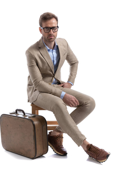atraente empresário posando em um chiar, descansando as mãos e vestindo óculos perto de uma pasta - Foto, Imagem