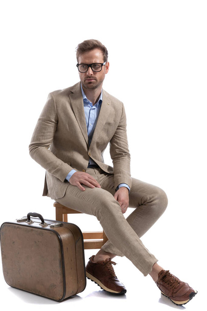 jeune homme d'affaires regardant ailleurs, assis sur un chiar, portant des lunettes près de sa mallette - Photo, image