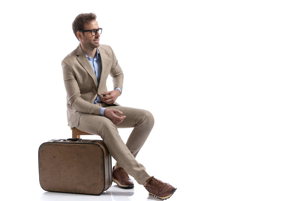 sexy businessman sitting on a chair, next to his briefcase, fixing his suit and wearing eyeglasses - Fotoğraf, Görsel